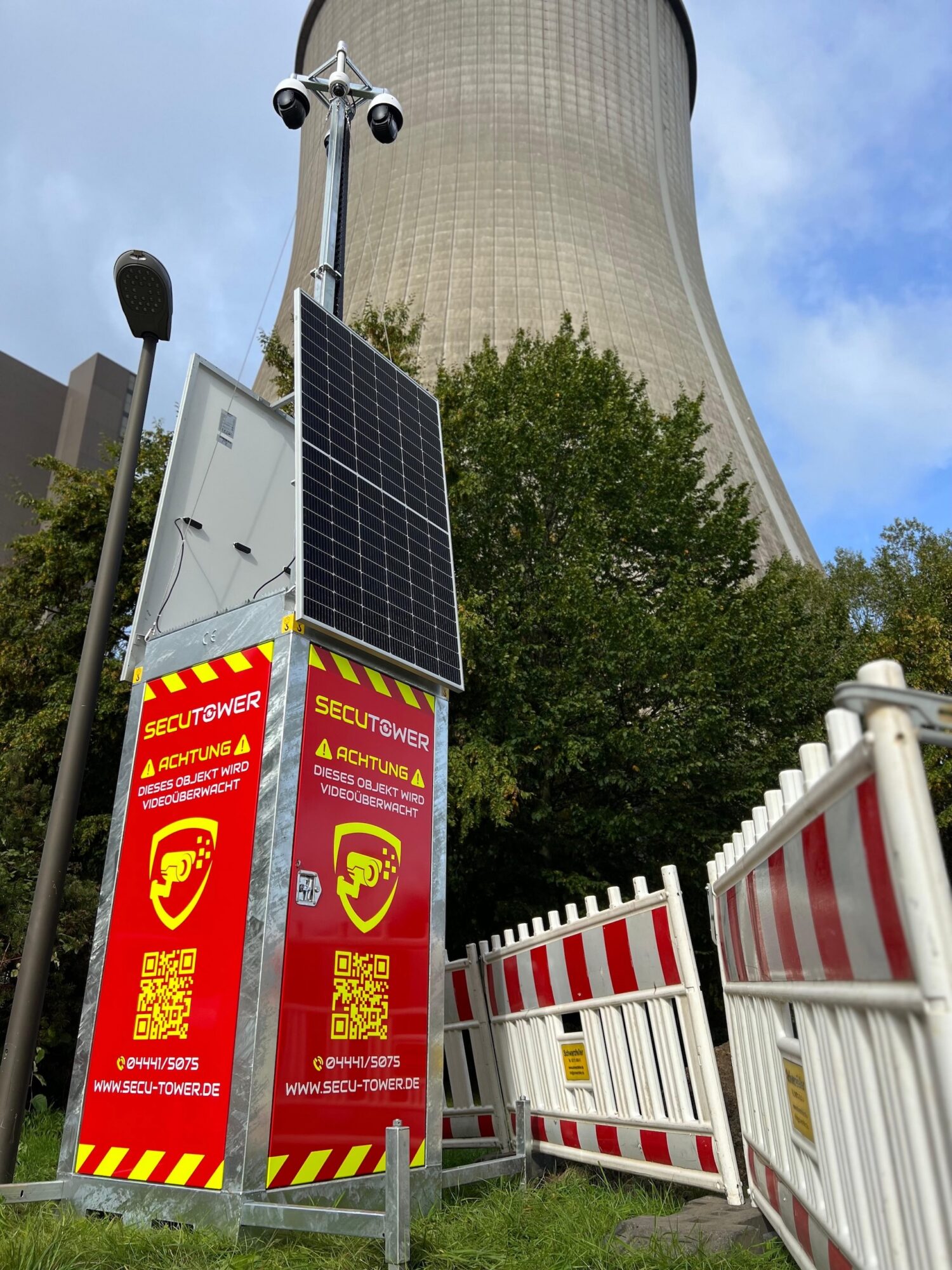 WhatsApp Image 2026-08-30 at 15.14.23 Mobile security tower with a solar panel and red warning signs beside a power plant cooling tower
